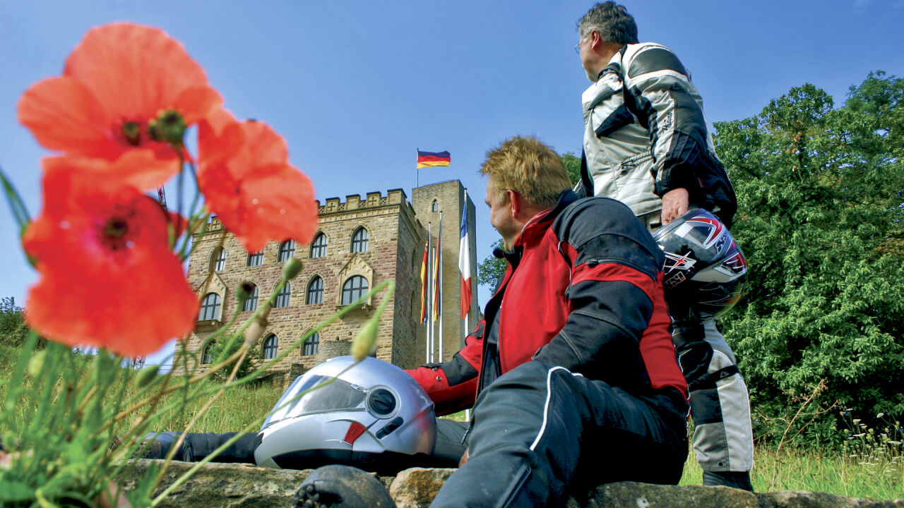 Zwei Motorradfahrer rasten vor einem Schloss mit Fahne, im Vordergrund Mohnblumen Zwei Motorradfahrer rasten vor einem Schloss mit Fahne, im Vordergrund Mohnblumen
